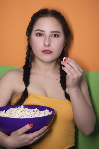charmante brunette vrouw in gele top met een paarse kom met popcorn op fel groene en oranje achtergrond. filmconcept. bioscoopthema - Foto, afbeelding
