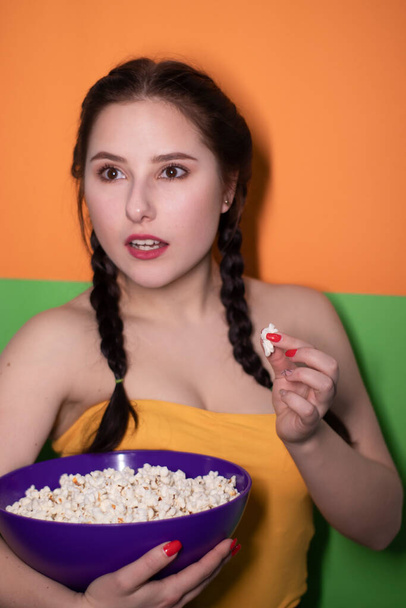 charming brunette woman in yellow top holding a purple bowl with popcorn on bright green and orange background. movie concept. cinema theme - Photo, image