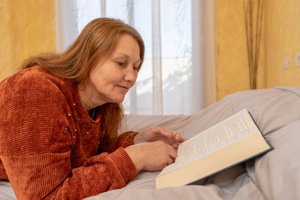 Mujer bastante adulta leyendo un libro sentado en la cama de su habitación. Concepto de entretenimiento. Enfoque selectivo - Foto, imagen