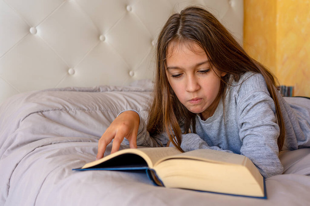 Jolie petite fille lisant un livre allongé sur le lit dans sa chambre. Concentration sélective. Concept d'apprentissage - Photo, image