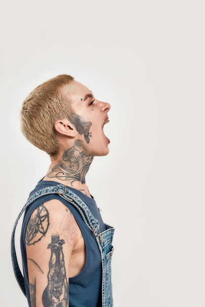 Emotions and postures. A white pierced and tattooed woman standing sideways to a camera wearing a denim overall screaming outloud with her mouth widely open - Foto, immagini