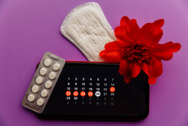 Menstruation app in smartphone with sanitary pad, pills and red flower on a lilac background. Woman critical days and hygiene protection concept - Photo, Image