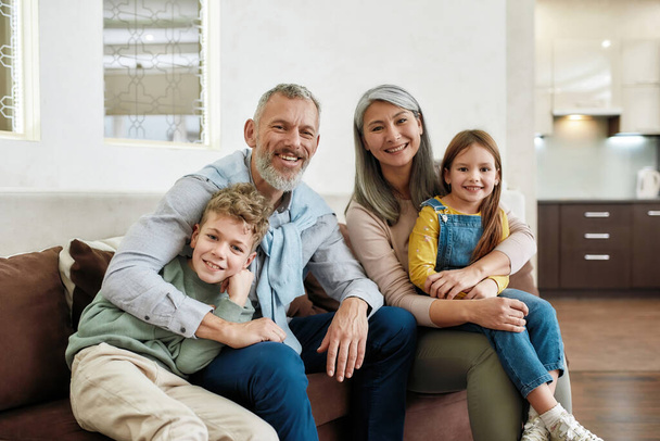 Portrait of happy family spending time at home, grandparents and grandchildren relaxing on sofa in the living room, smiling at camera - 写真・画像