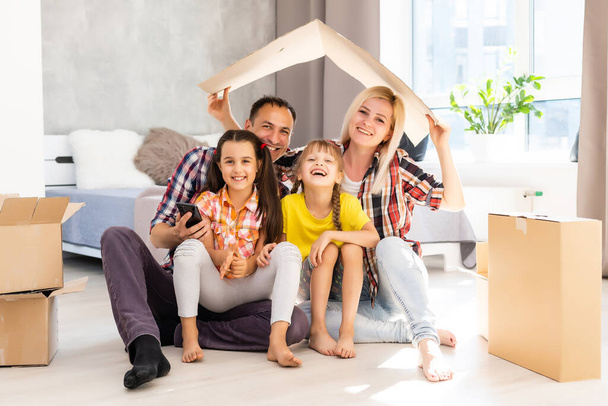 Caucasian family, man, woman and two girls sit on floor, unpack boxes and smile in new house. Behind them moving boxes. - Photo, Image