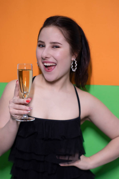 okouzlující brunetka žena v černých šatech drží sklenici šampaňského s zásnubním prstenem uvnitř na jasně zeleném a oranžovém pozadí. svatební koncept. Zasnoubení. a ptám se. nevěsta - Fotografie, Obrázek