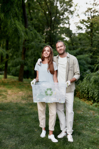 Vertical shot of young happy couple, eco volunteers holding recycle bin and looking at camera while collecting plastic waste together in the green forest - Фото, изображение