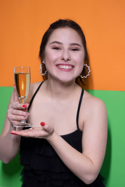 charming brunette woman in black dress holding glass of champagne with an engagement ring inside on bright green and orange background. wedding concept. engagement. poping the question. bride - Фото, изображение
