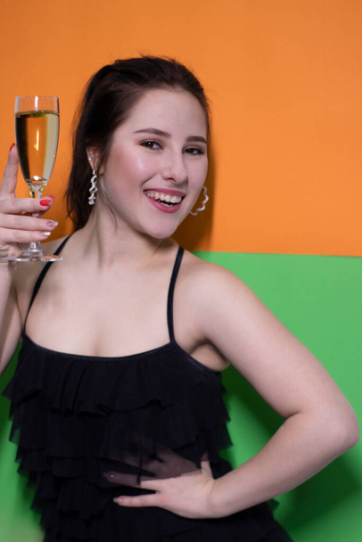 очаровательная брюнетка в черном платье держит бокал шампанского с обручальным кольцом внутри на ярко-зеленом и оранжевом фоне. свадебная концепция. Помолвка. задаю вопрос. невеста - Фото, изображение
