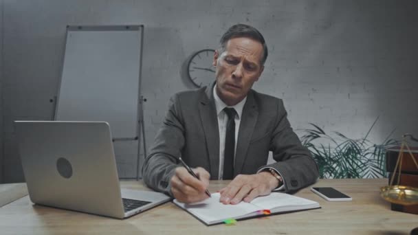 Droevige verzekeringsagent schrijven op notebook in de buurt van apparaten in het kantoor  - Video