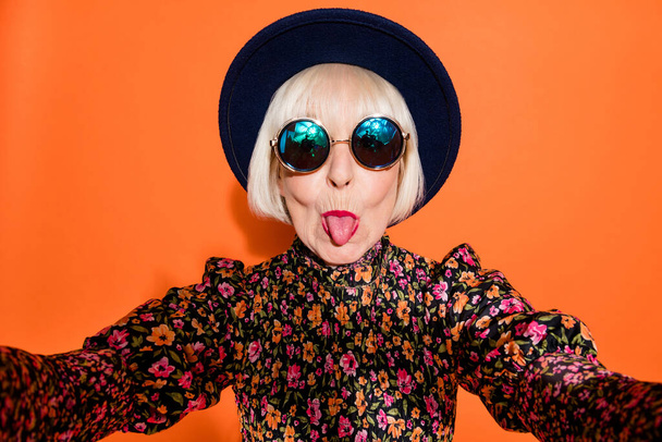 Portrait of old funky cool crazy hipster grandmother take selfie stick tongue out isolated on orange color background - Photo, Image