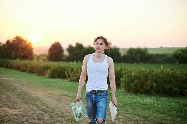 Young male artist, wearing torn jeans and white t-shirt, walking on green field during sunset, holding canvas and palette thinking. Painting workshop in countryside. Artistic education concept. - Valokuva, kuva