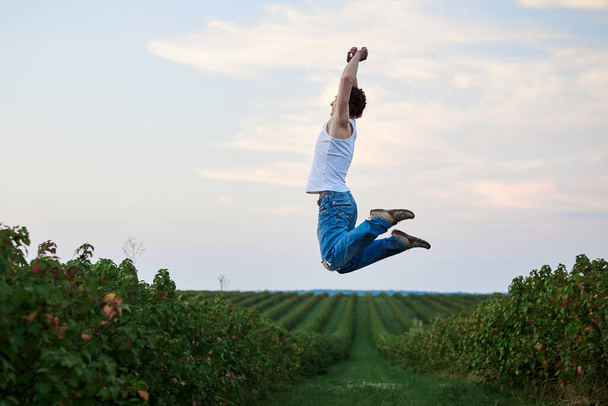 Young handsome man, wearing torn jeans and white t-shirt, jumping in the air on green field, smiling, laughing, having fun. Artistic education concept. Creative portrait outside. Freedom inspiration. - Photo, image