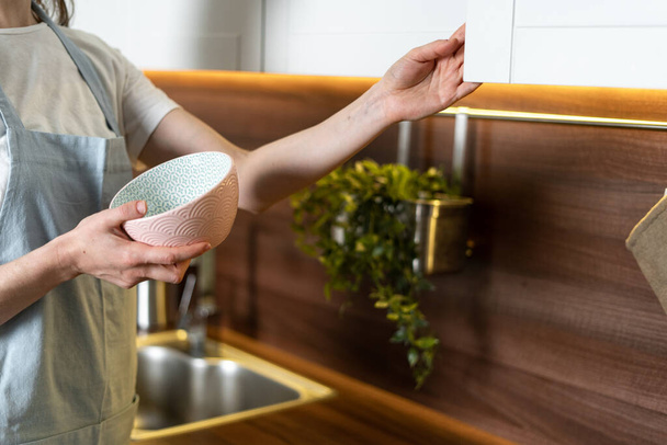 Cropped view of woman in blue apron standing on modern kitchen, open door and puts clean ceramic dishes in the cupboard. Housewife making daily routine stuff concept - Фото, изображение