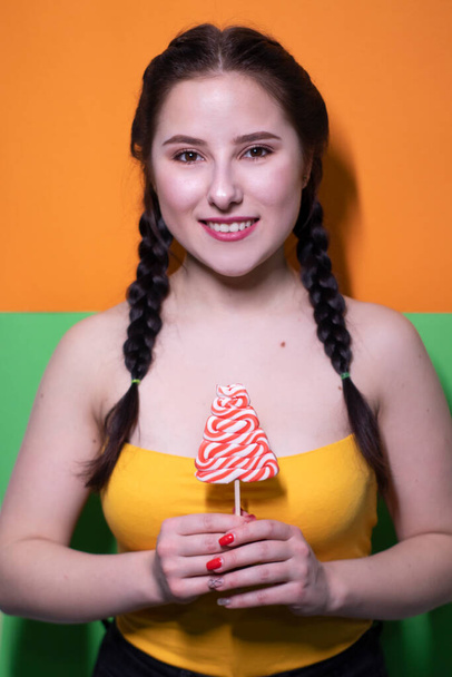 charming brunette woman in yellow top holding red big lollipop on bright green and orange background. sweets concept. candy. copy space - Foto, imagen