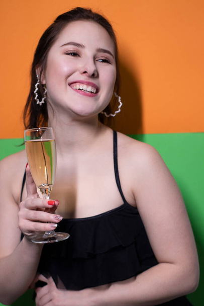 Charmante brünette Frau im schwarzen Kleid mit einem Glas Champagner und einem Verlobungsring drinnen auf leuchtend grünem und orangefarbenem Hintergrund. Hochzeitskonzept. Engagement. Die Frage drängt sich auf. Braut - Foto, Bild