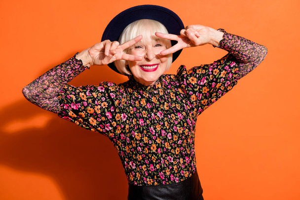 Fotoporträt von lustigen fröhlichen Seniorin zeigt V-Zeichen Geste trägt schwarze Kleidung isoliert auf lebendigen orangefarbenen Hintergrund - Foto, Bild