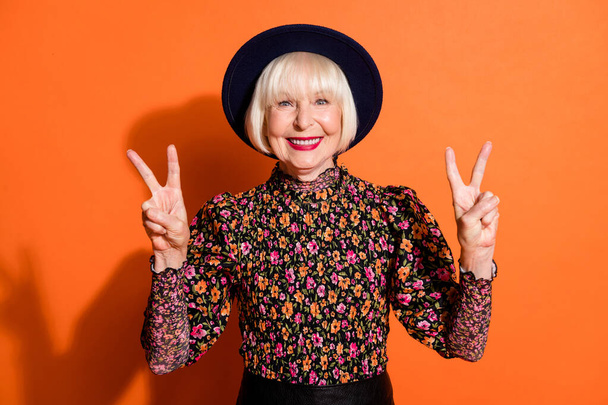 Photo portrait of cheerful happy stylish senior woman showing v-sign peace gesture smiling isolated on vibrant orange background - Photo, image