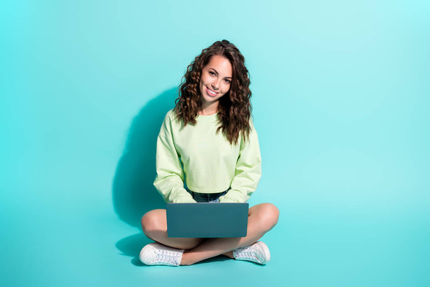 Photo of charming young girl sit floor hold laptop beaming smiling look camera wear green sweater sneakers isolated blue color background - Fotó, kép