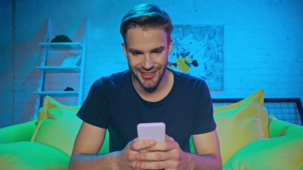 Sorrindo homem usando smartphone no sofá durante a noite  - Filmagem, Vídeo