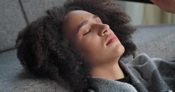 Портрет сумної афро-американської жінки етнічної змішаної раси, яка лежить на дивані, думаючи про проблеми, що утримують її лоб руками, відчувають мігрені симптоми хвороби і стрес концепції - Кадри, відео
