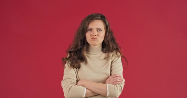 Young angry offended girl fashionable model sad girlfriend stands on red background demonstrates displeasure jealousy folds hands turns away frowning female face, concept of problems and quarrels - Footage, Video