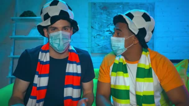 Multikulturelle Fußballfans in medizinischen Masken mit Bier auf der Couch  - Filmmaterial, Video