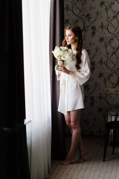 beautiful woman in a white robe with a wedding bouquet of flowers at the window - Фото, зображення