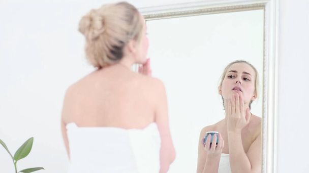 Señora mirando en el espejo, la aplicación de crema de loción hidratante en las mejillas, terminando rutina de cuidado de la piel doméstica por la mañana. Mujer sonriente arreglándose después de ducharse en el baño. - Foto, imagen