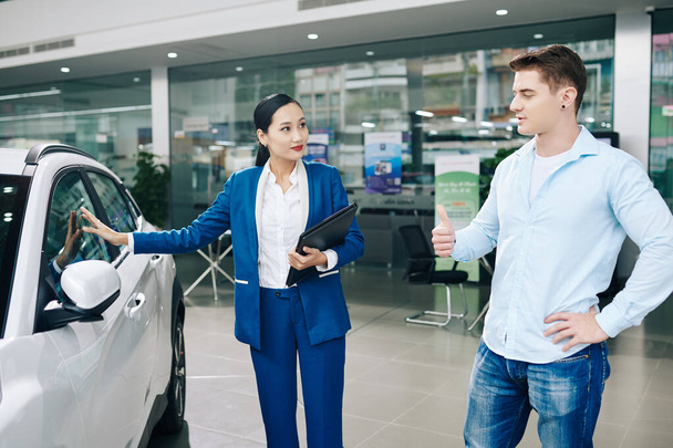 Pretty young manager showing automobile to smiling young car dealership client - Photo, Image