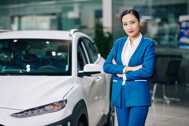Porträt der hübschen jungen Autohausmanagerin, die neben dem Auto steht, die Arme verschränkt und in die Kamera blickt - Foto, Bild