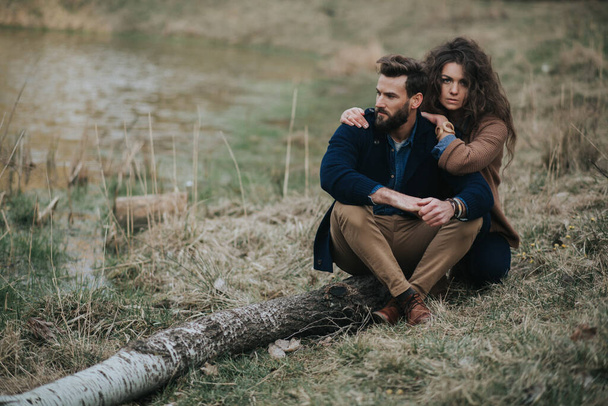 Χαρούμενοι λευκοί εραστές κάθονται στην όχθη της λίμνης. Νεαρό ζευγάρι αγκαλιάζεται την ημέρα του φθινοπώρου έξω. Ένας γενειοφόρος άντρας και μια σγουρή γυναίκα ερωτευμένη. Του Αγίου Βαλεντίνου. Έννοια της αγάπης και της οικογένειας - Φωτογραφία, εικόνα