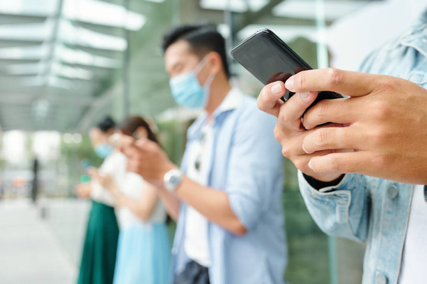 Detailní záběr mladých lidí stojících venku v lékařských maskách a textujících si navzájem, zaměřený na popředí - Fotografie, Obrázek