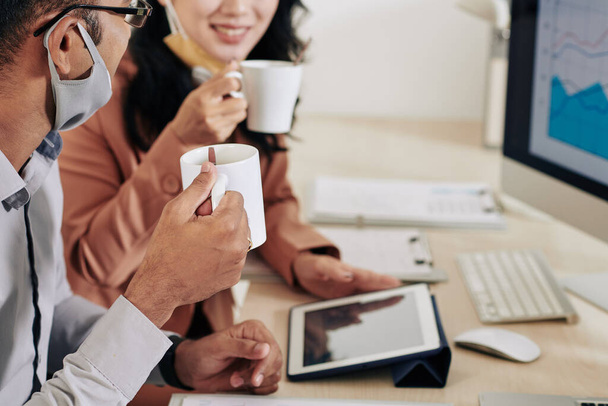 Immagine ritagliata di colleghi d'affari sorridenti che bevono caffè e discutono rapporti e grafici alla riunione in ufficio - Foto, immagini