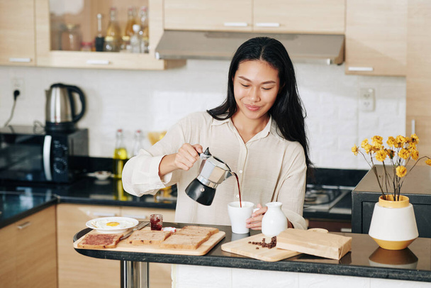 Sonriendo bonita mujer china vertiendo café en taza cuando desayuna en la cocina - Foto, Imagen