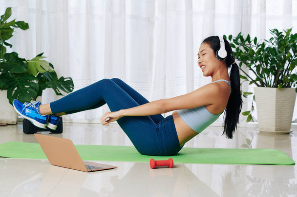 Cheerful young woman in headphones repeating after online trainer when doing abs exercise - Photo, Image