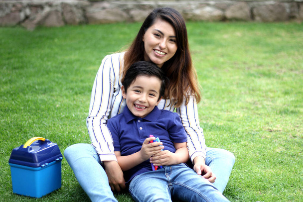 Latino maman et 6 ans garçon assis sur l'herbe avec boîte à lunch et couleurs pour la rentrée scolaire - Photo, image