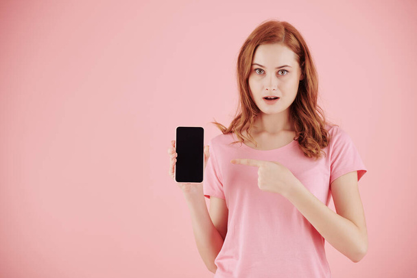 Ritratto di giovane donna scioccata con i capelli rossi che puntano sullo schermo dello smartphone - Foto, immagini