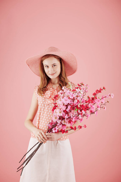 Portrait of smiling young pretty woman in straw hat holding branches of blooming oriental cherry - Foto, afbeelding