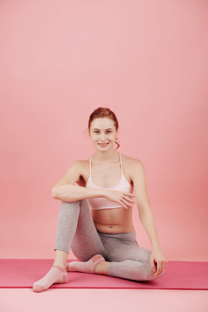 Mujer joven bastante sonriente descansando en la esterilla de yoga después de hacer ejercicio en el estudio rosa - Foto, Imagen