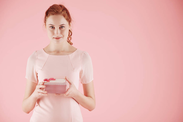 Portrait of smiling young pretty red haired woman with gift box standing against pink background - Photo, image
