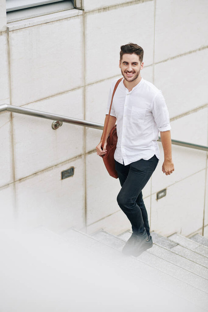Веселий красивий молодий бородатий чоловік у бавовняній білій сорочці, що йде по сходах
 - Фото, зображення