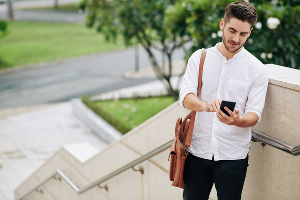 Schöner junger Mann in weißem Hemd, der auf der Treppe steht und SMS von Freund oder Kollege beantwortet - Foto, Bild