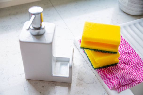 Two yellow sponges, wiping cloth and liquid dispenser on a marble kitchen counter. - Photo, image
