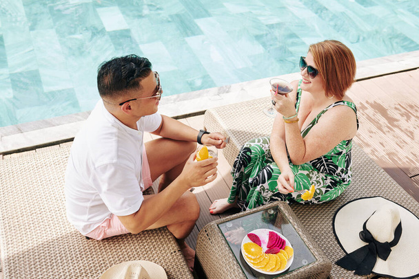 Marido e mulher sorridentes bebendo coquetéis, comendo frutas e conversando ao descansar na piscina - Foto, Imagem