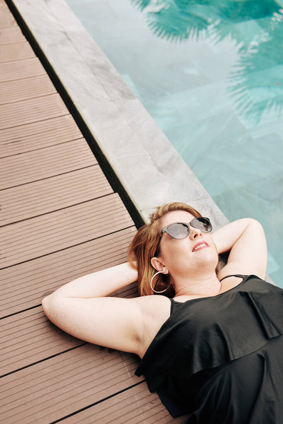 улыбающаяся женщина в солнечных очках, загорающая у бассейна спа-отеля, вид сверху - Фото, изображение