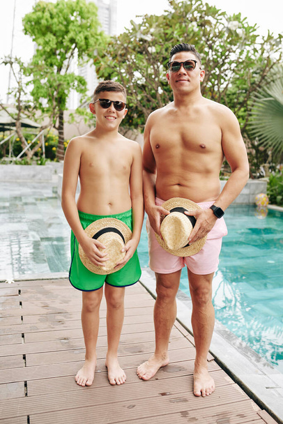Mosolygó, izmos férfi napszemüvegben, és a kisfia, aki a medence mellett áll, kalapokkal a kezében. - Fotó, kép