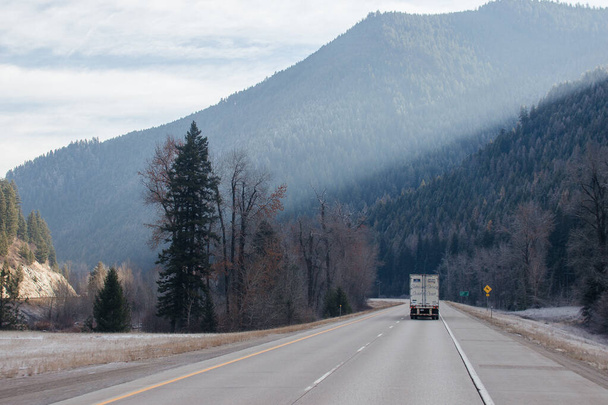 Asfalttitie, jota pitkin autot ja kuorma-autot kulkevat vuorten välissä, eteenpäin horisontissa vuorilla sumussa, taivas on pilvien peitossa. Idaho, Yhdysvallat, 11-23-2019 - Valokuva, kuva