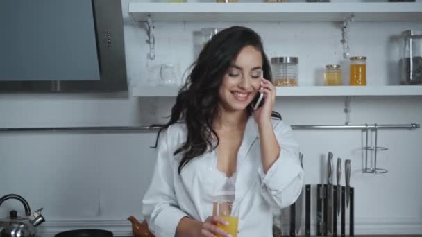 χαρούμενη, σέξι γυναίκα κρατώντας χυμό πορτοκαλιού και μιλώντας στο smartphone στην κουζίνα - Πλάνα, βίντεο