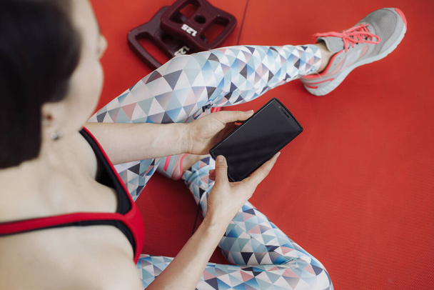 Young woman resting after workout. Healthy young female taking break after training in gym with phone in hands. - Photo, image