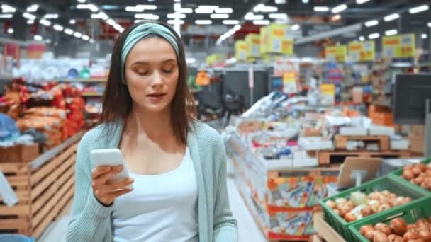 nuori nainen tarkistaa listan älypuhelimeen kävellessään supermarketissa - Materiaali, video